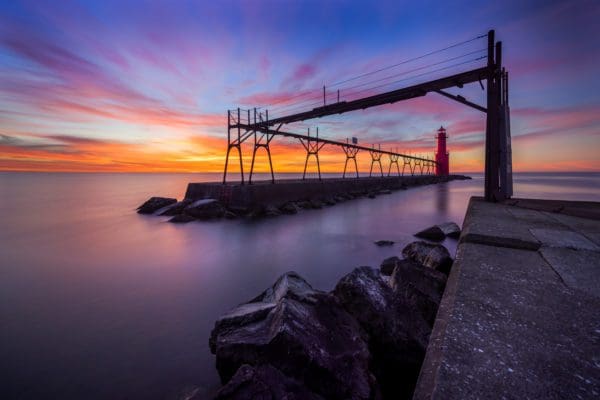 A multi-colored sunset highlights a red lighthouse and the maintenance framework that leads to it.