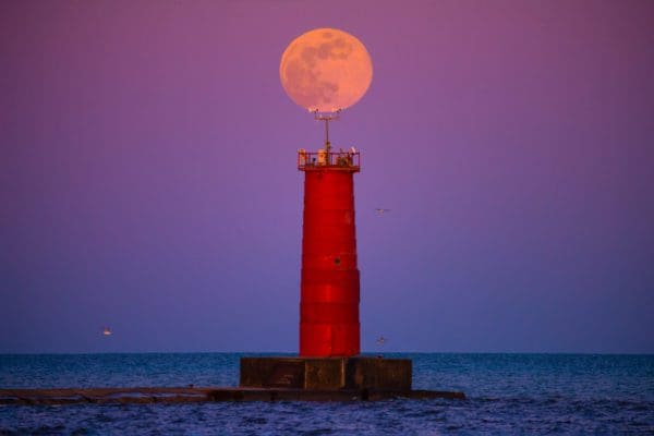 A red lighthouse with a full moon perfectly centered above it.
